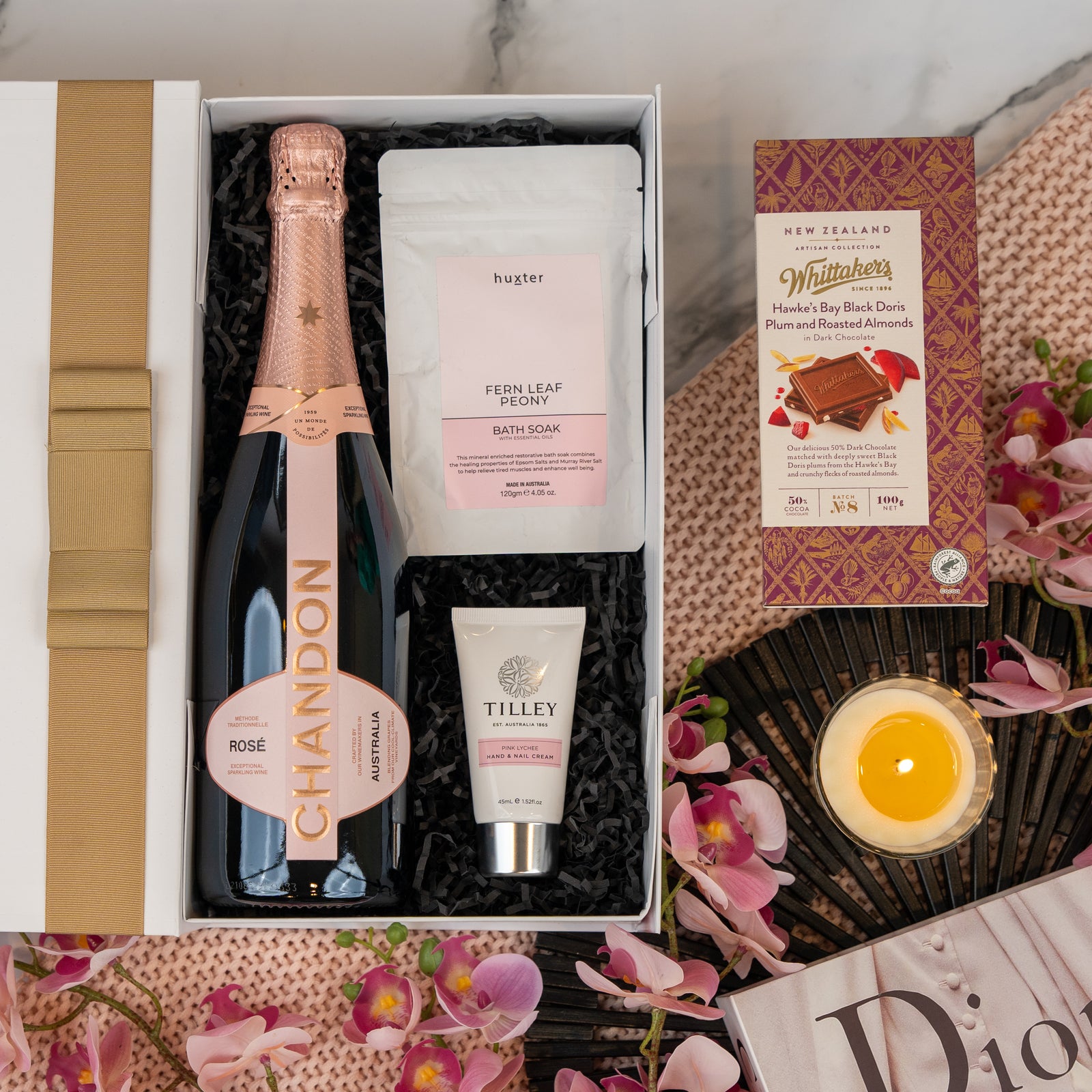The Hamper Boutique Co’s Chandon Rosé Indulgence Hamper includes Chandon rosé, Huxter bath soak, Tilley hand cream, and Whittakers plum chocolate, styled with pink flowers, a yellow candle, and a Dior booklet on marble.