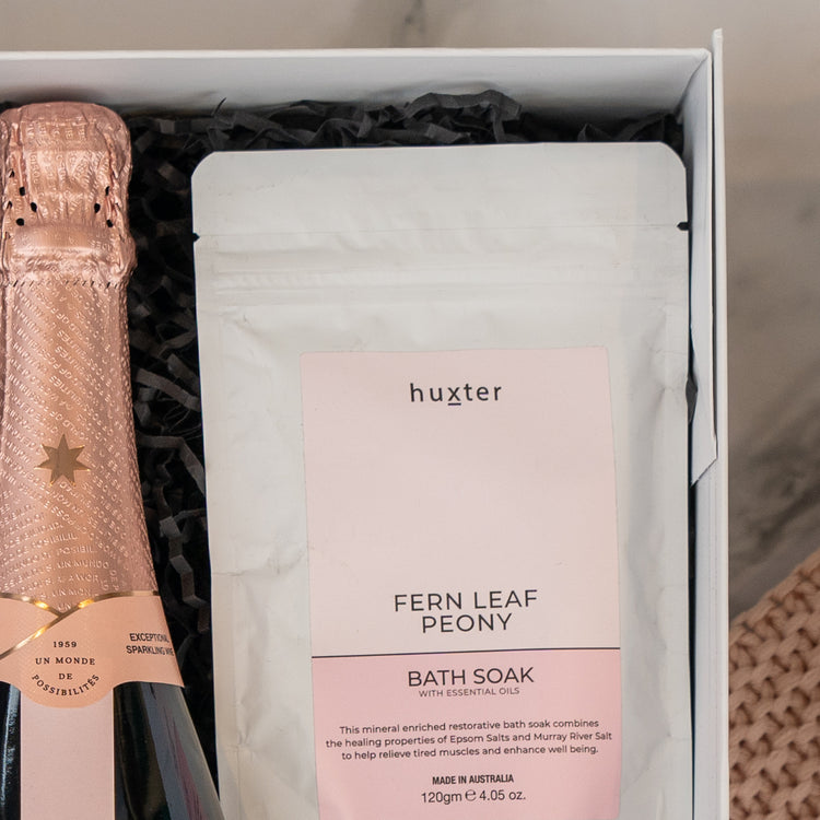The Chandon Rosé Indulgence Hamper by The Hamper Boutique Co includes a sparkling wine with pink foil and Huxter Fern Leaf Peony bath soak, all elegantly presented in black shredded paper.