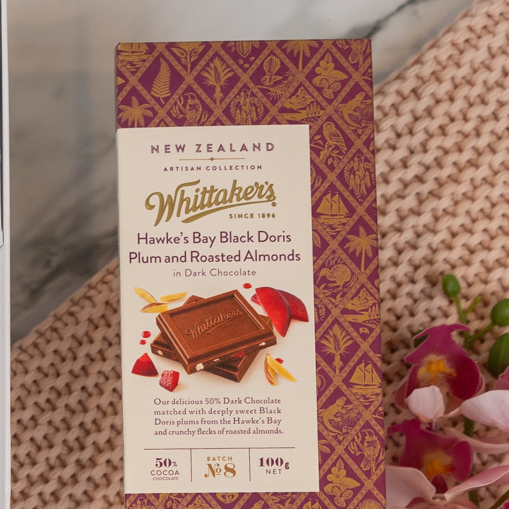 A Chandon Rosé Indulgence Hamper by The Hamper Boutique Co sits on a woven mat, with pink orchid flowers beside it. The hamper box features a purple and gold design.