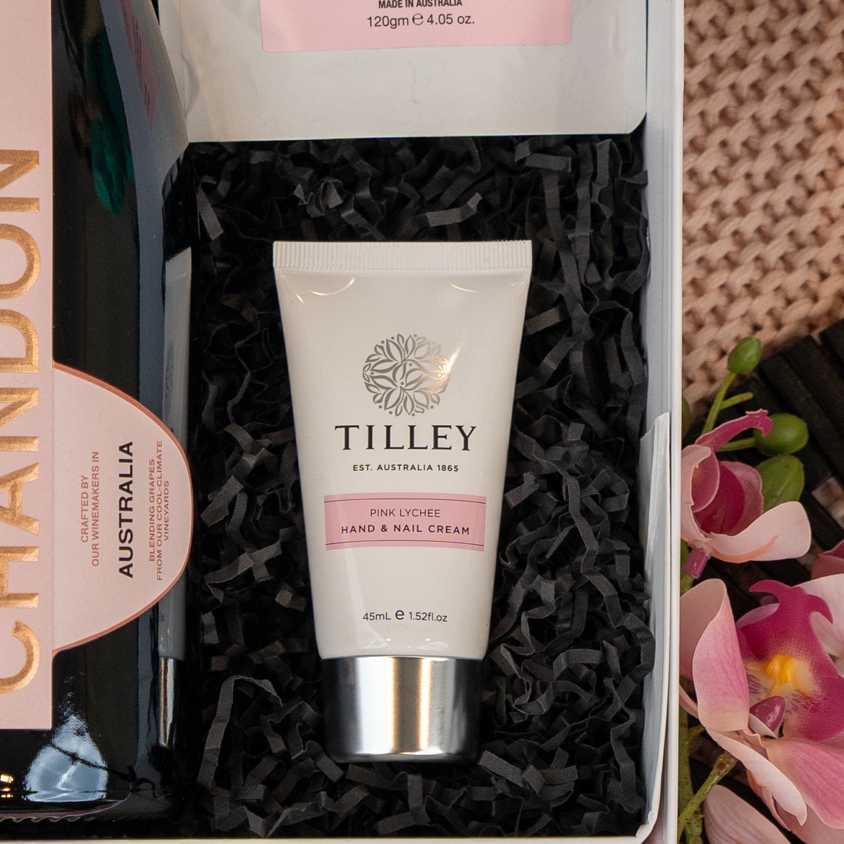 The Hamper Boutique Co's Chandon Rosé Indulgence Hamper includes a tube of Tilley Pink Lychee Hand & Nail Cream in a gift box with black shredded paper, paired with Chandon sparkling wine and a pink orchid flower.