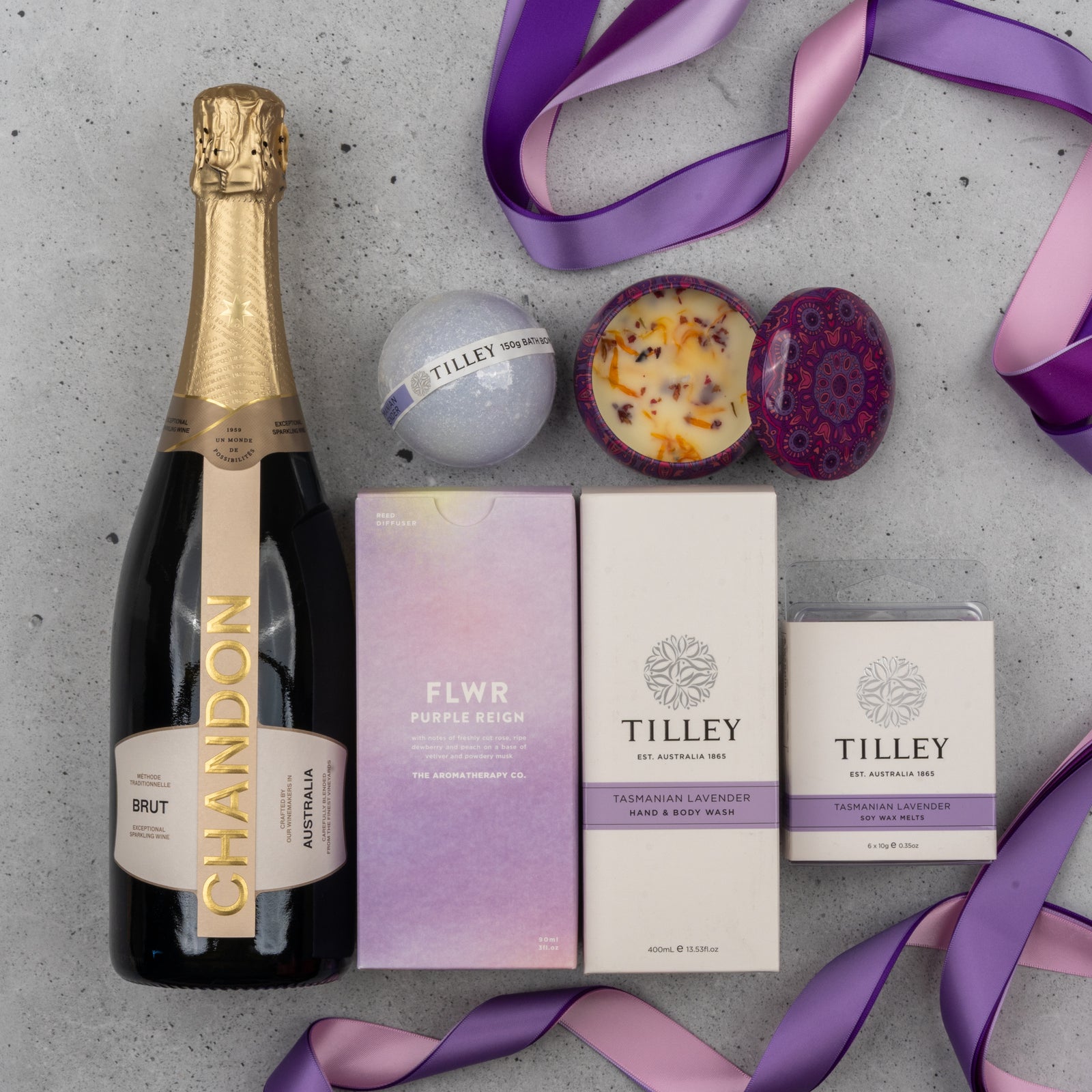 The Chandon Deluxe Celebration Hamper by The Hamper Boutique Co features Chandon sparkling wine, a purple bath bomb, a floral candle, and two Tilley Tasmanian Lavender hand and body wash products, tied with purple and pink ribbon on a gray surface.
