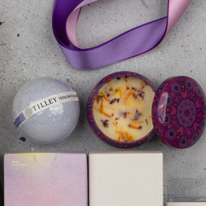 The Chandon Deluxe Celebration Hamper by The Hamper Boutique Co features a purple tin candle with dried flowers, a Tilley bath bomb, a purple and pink ribbon, and pastel boxes, all beautifully arranged on a gray surface.