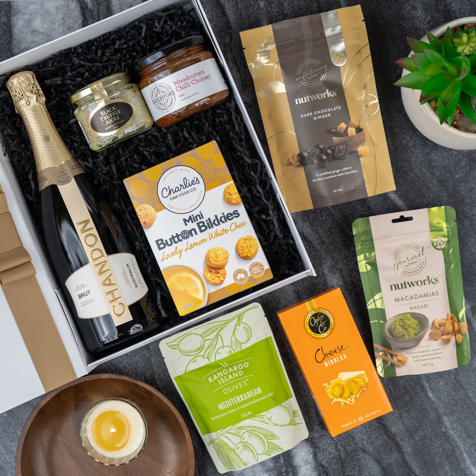 The Chandon Gourmet Sparkling Gift Hamper by The Hamper Boutique Co features sparkling wine, jam, chutney, lemon white choc biscuits, cheese sticks, olives, wasabi and dark choc macadamias, plus a lit candle on a marble surface.