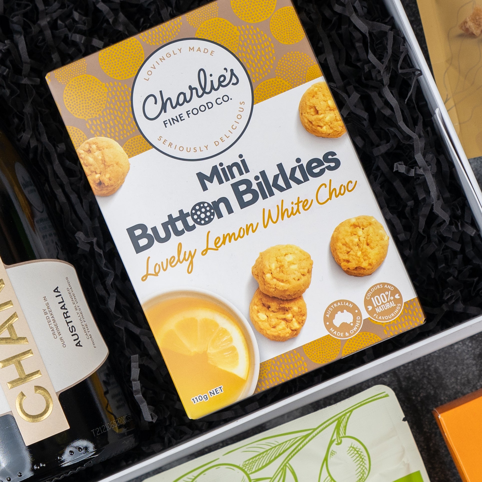 A Chandon Gourmet Sparkling Gift Hamper by The Hamper Boutique Co includes Charlie’s Fine Food Co. Mini Button Bikkies Lovely Lemon White Choc, with black shredded paper, a bottle labeled Australia, and another packaged item nearby.