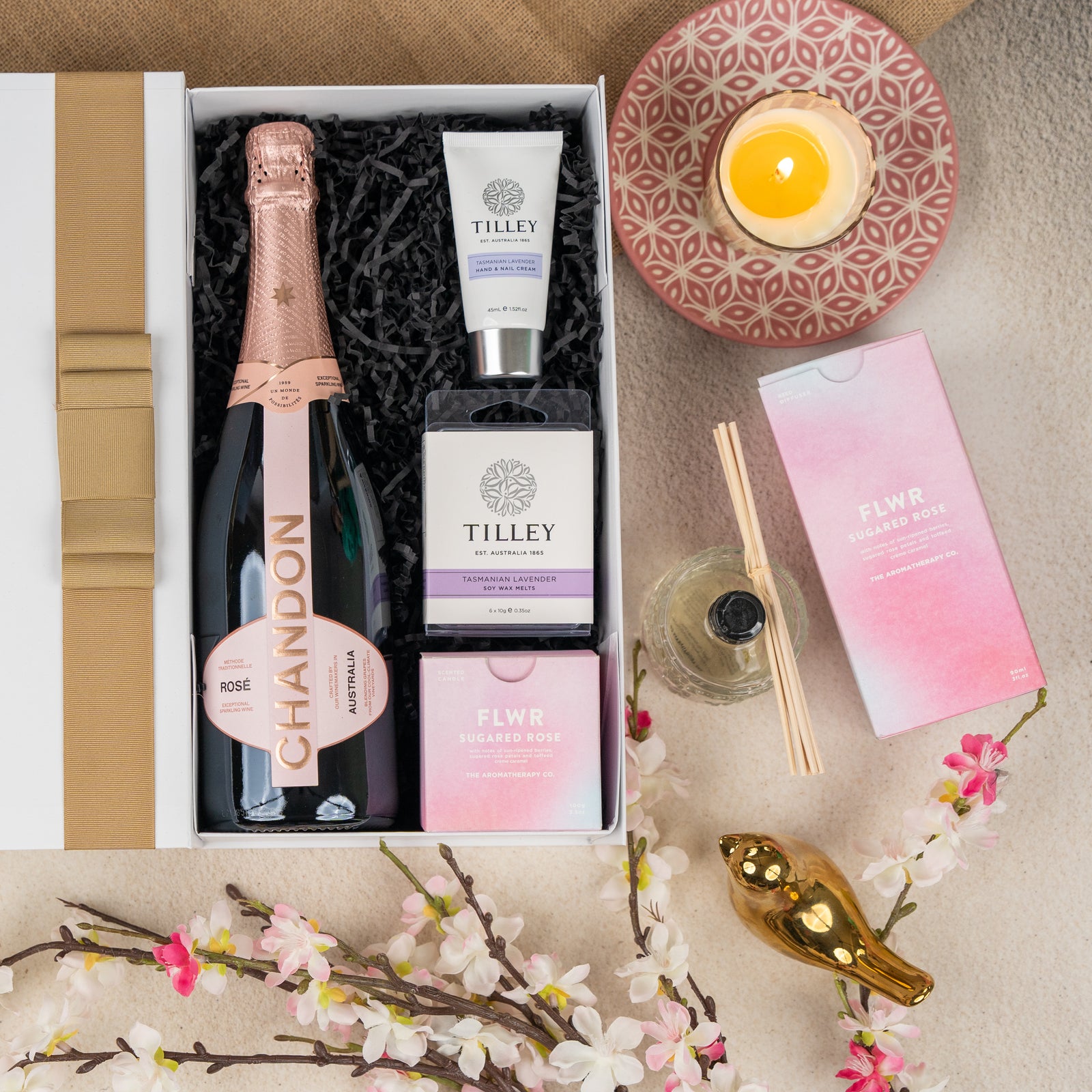 The Hamper Boutique Co's Chandon Bloom Retreat gift box features Chandon rosé, Tilley hand cream and soap, a Sugared Rose reed diffuser, yellow candle, pink floral accents, and a gold bird ornament arranged on a beige surface.