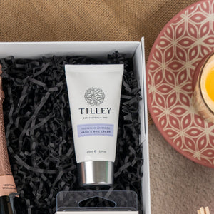 A Chandon Bloom Retreat gift box by The Hamper Boutique Co sits with black shredded paper next to a glass of orange juice on a pink patterned plate.