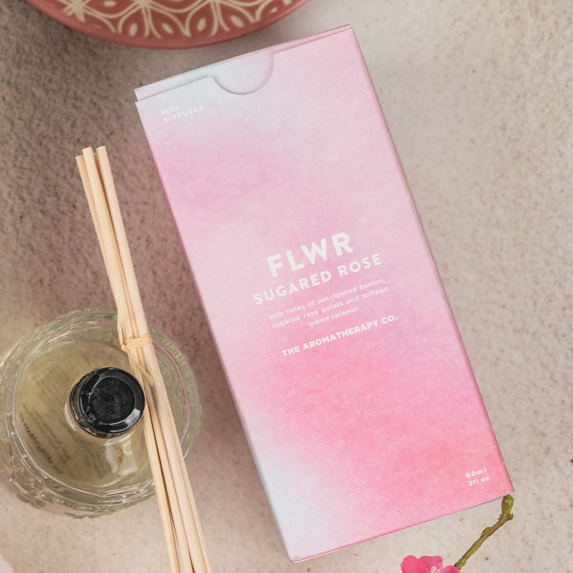 A box labeled Chandon Bloom Retreat by The Hamper Boutique Co sits next to a glass diffuser bottle with reed sticks on a light surface, while part of a floral decoration is visible in the corner.