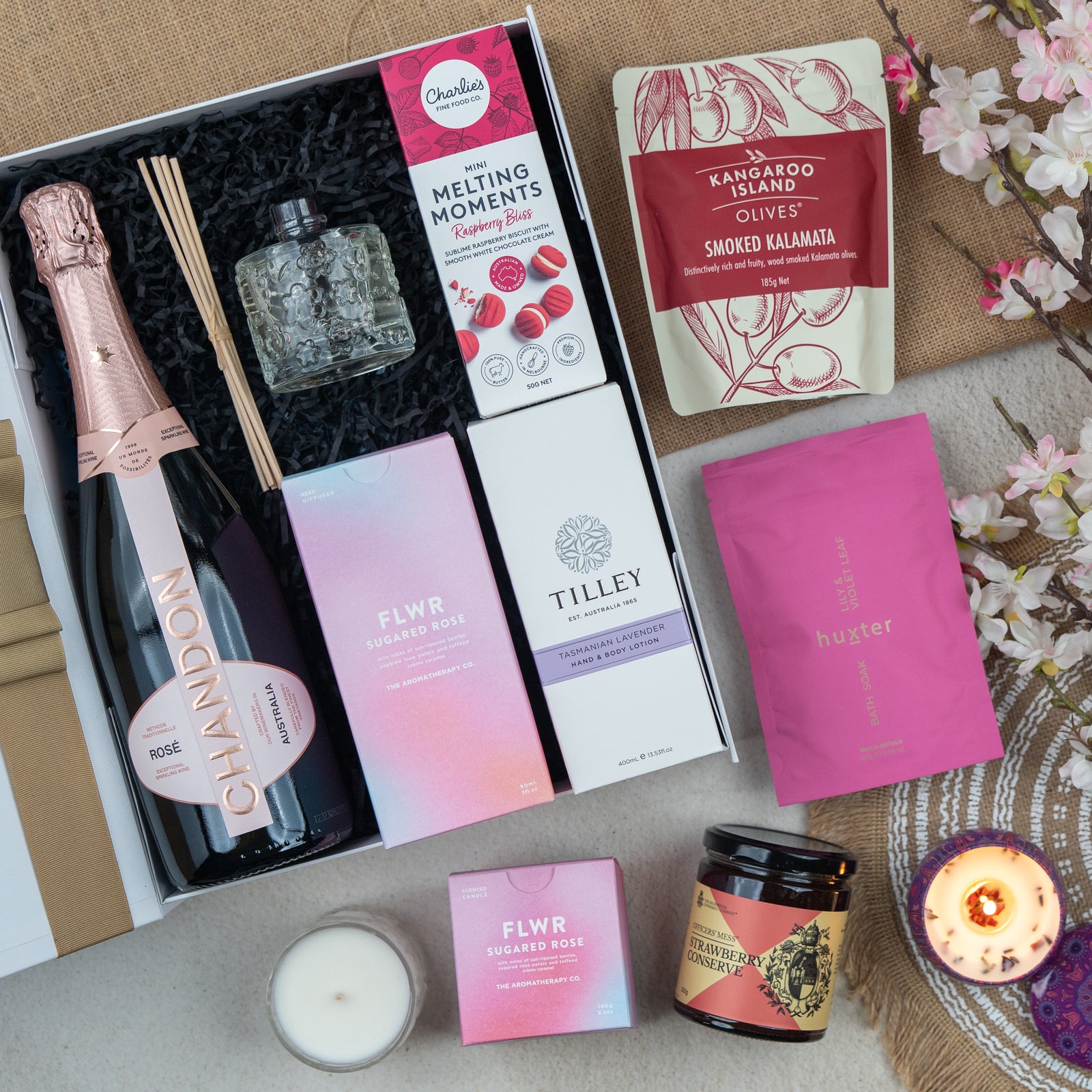 A flat lay of The Hamper Boutique Co’s Chandon Connoisseur's Selection Hamper with Chandon rosé, diffuser, candle, floral soap, raspberry bliss cookies, olives, sugared rose chocolates, pink pouch, honey jar. Flowers and a tealight are nearby.