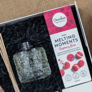 The Hamper Boutique Co's Chandon Connoisseur's Selection Hamper features a floral glass diffuser bottle and Charlie’s Fine Food Co. Mini Melting Moments Raspberry Bliss biscuits, all elegantly presented on black shredded paper.