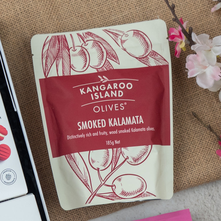 A Chandon Connoisseur's Selection Hamper by The Hamper Boutique Co sits on a textured surface next to a branch with white and pink flowers. The package features elegant cream coloring with red illustrations.