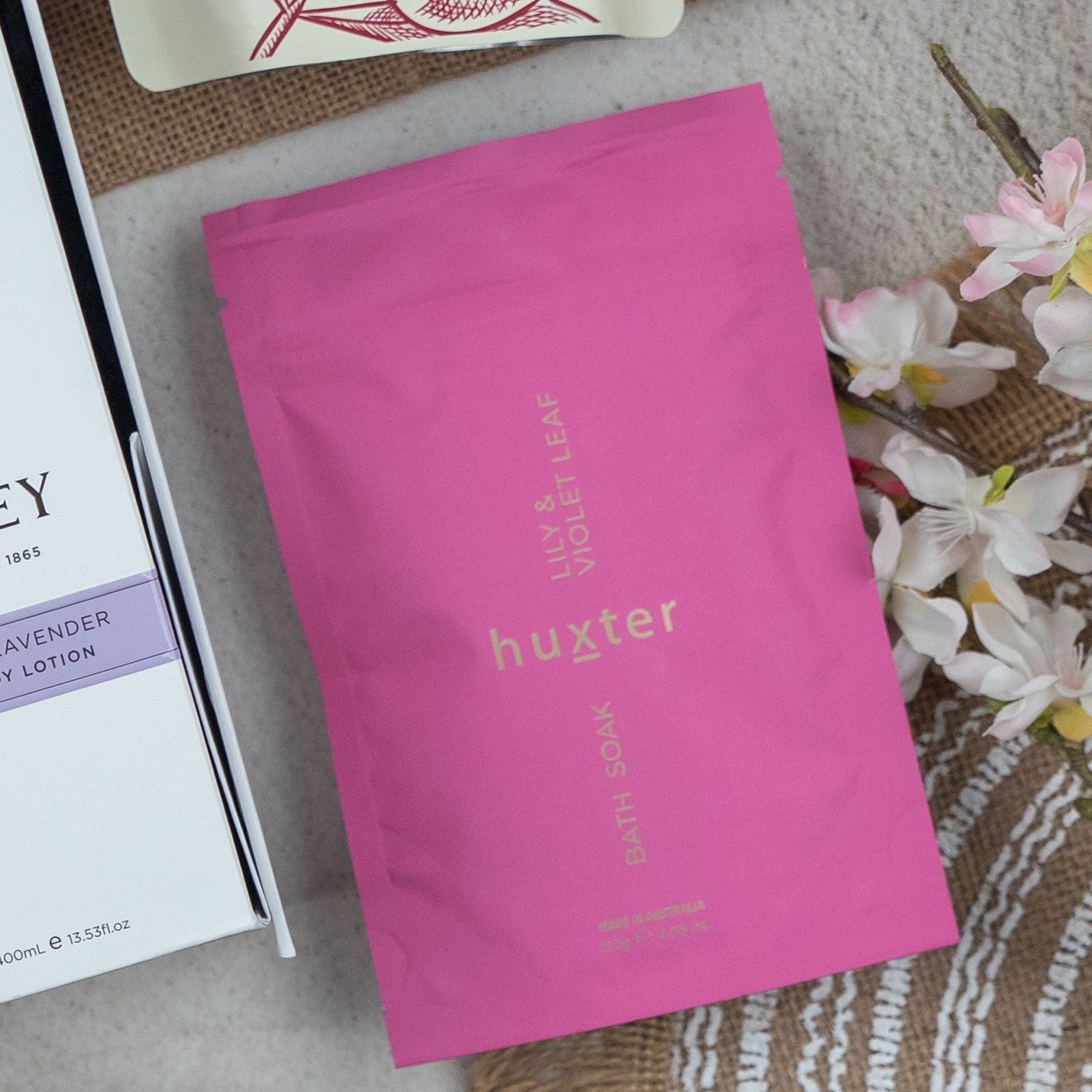 A Chandon Connoisseur's Selection Hamper from The Hamper Boutique Co is displayed on a beige surface beside white and pink artificial flowers, a white box, and part of a woven basket.
