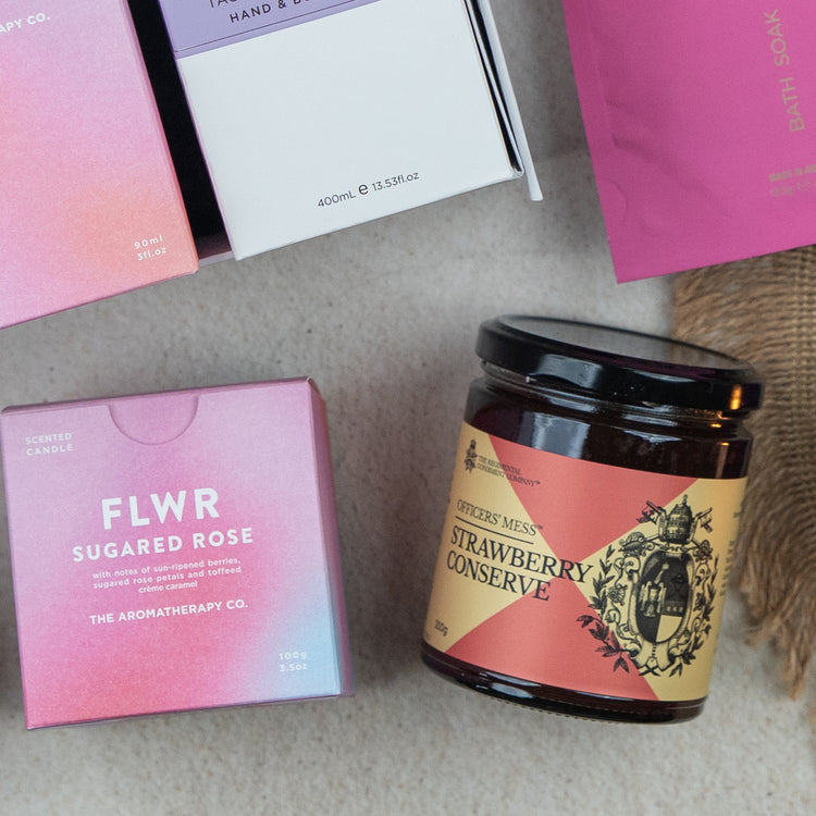 The Hamper Boutique Co's Chandon Connoisseur's Selection Hamper features a pink scented candle box, Officers Mess Strawberry Conserve jar, a white bottle, and pink bath soak packaging arranged on a light surface.