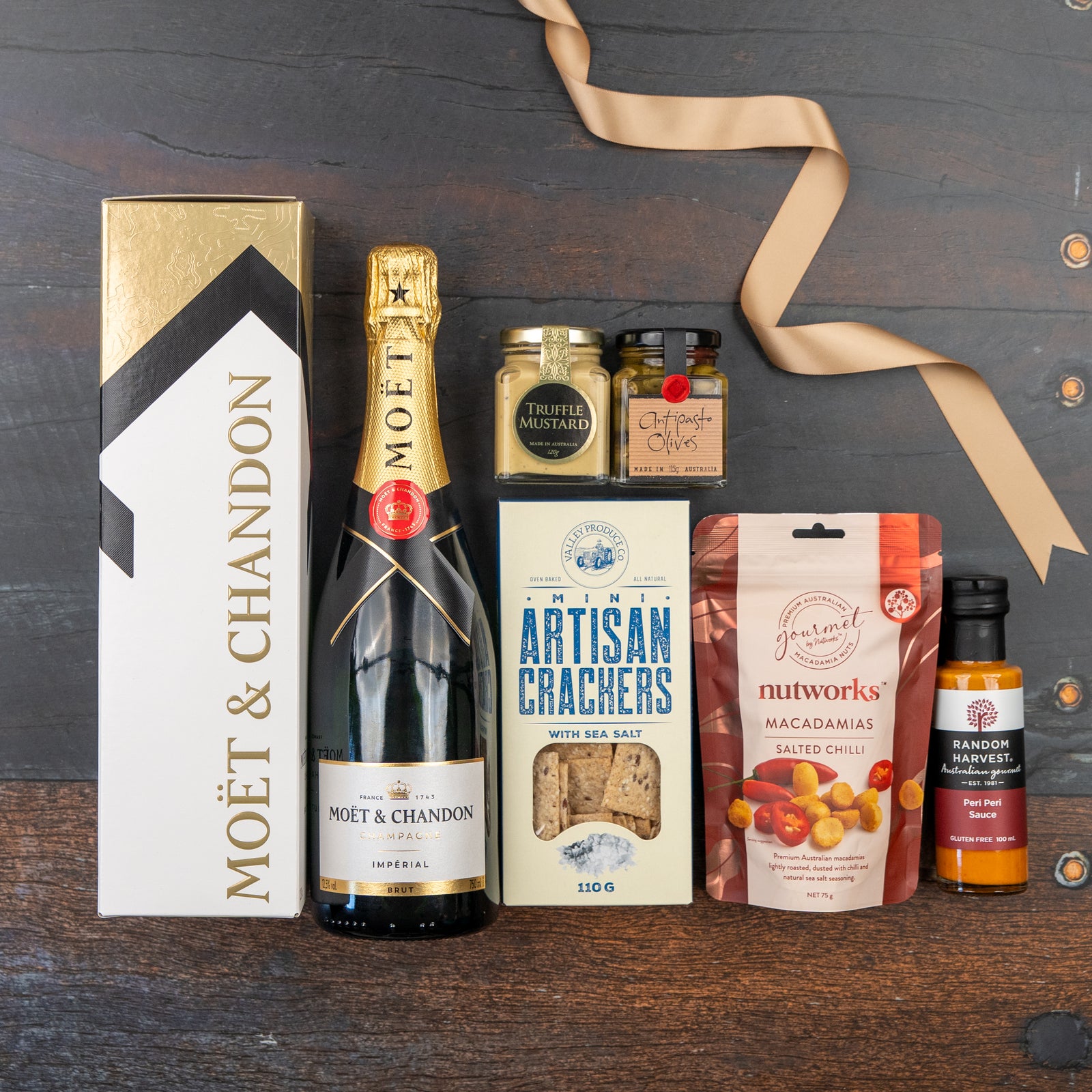 The Hamper Boutique Co's Moët & Chandon Celebration Hamper sits on a dark wooden surface, featuring Champagne, artisan crackers, truffle mustard, herb olives, salted chili macadamias, dressing, and a beige ribbon.