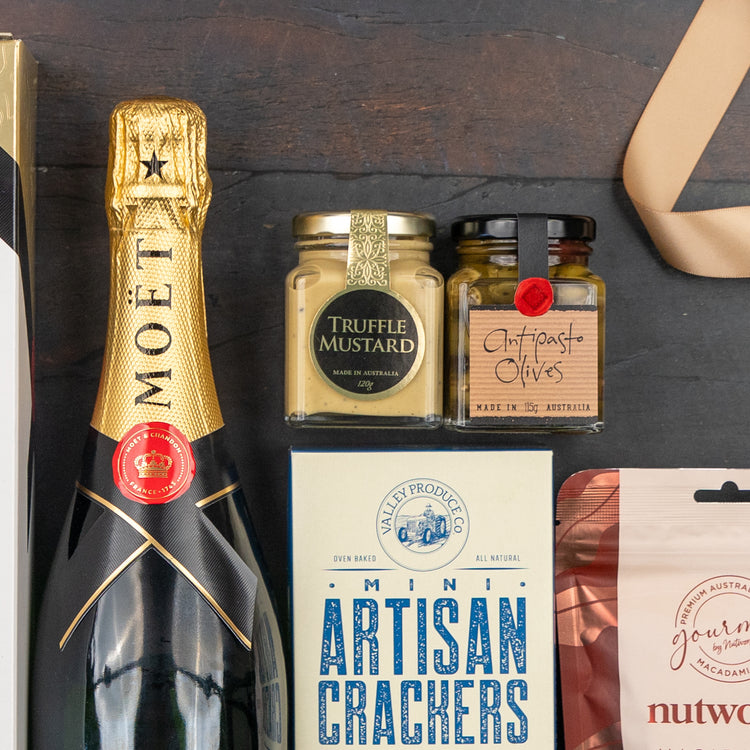 The Hamper Boutique Co's Moët & Chandon Celebration Hamper includes Moët champagne, truffle mustard, antipasto olives, mini artisan crackers, and a nut mix elegantly arranged on a dark surface.