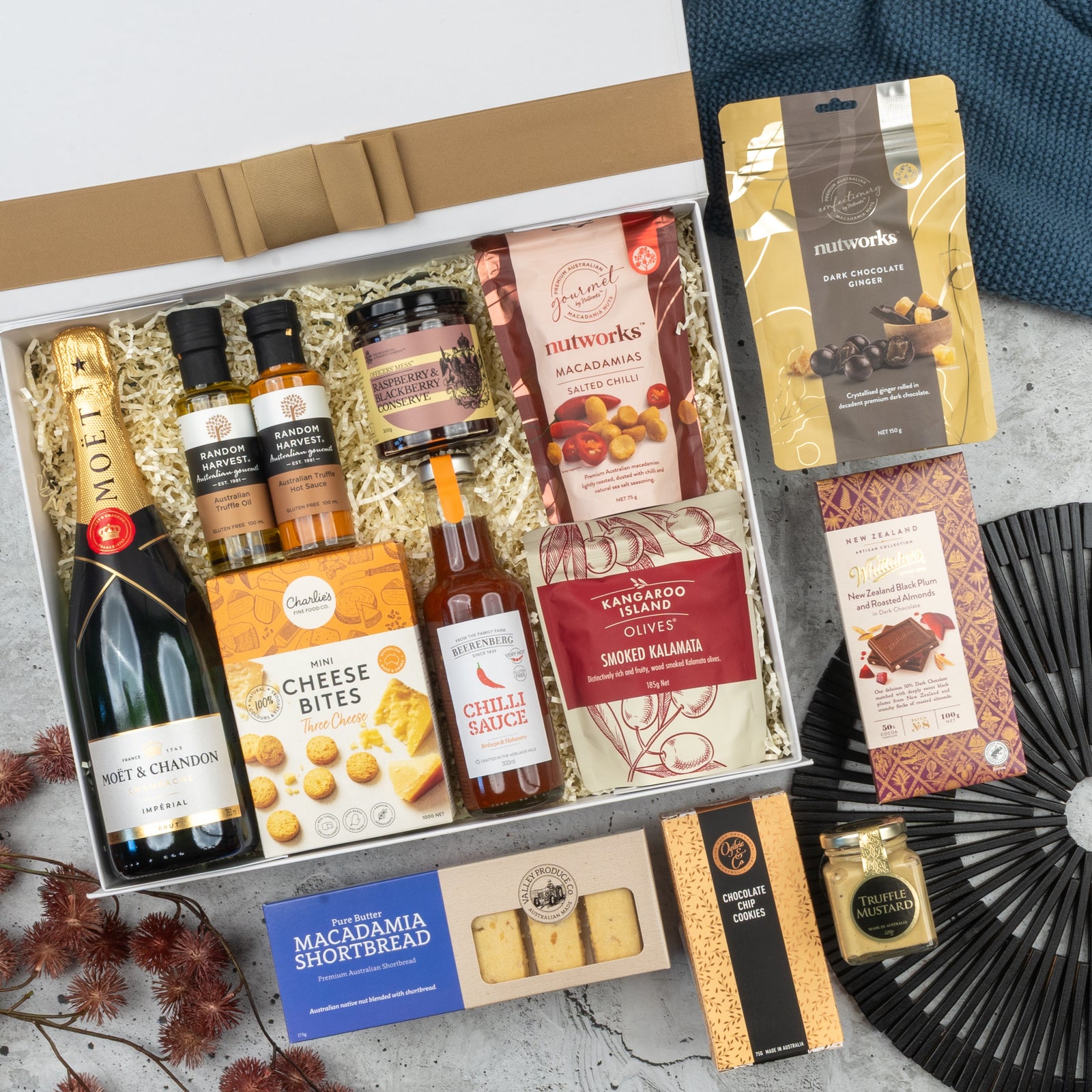 The Hamper Boutique Co’s Moët & Chandon Deluxe Celebration Hamper includes champagne, sauces, cheese bites, olives, macadamias, shortbread, chocolate, honey & snacks in a stylish white box on a grey surface.