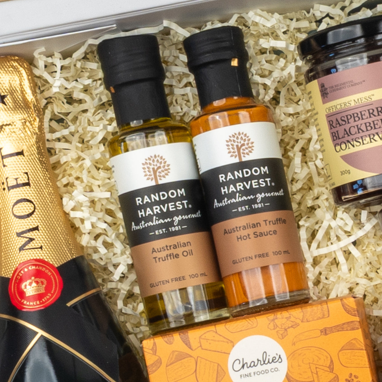 A close-up of The Hamper Boutique Co Moët & Chandon Deluxe Celebration Hamper filled with a bottle of Moët champagne, Random Harvest truffle products, raspberry blackberry conserve, and Charlies cookies on shredded paper.