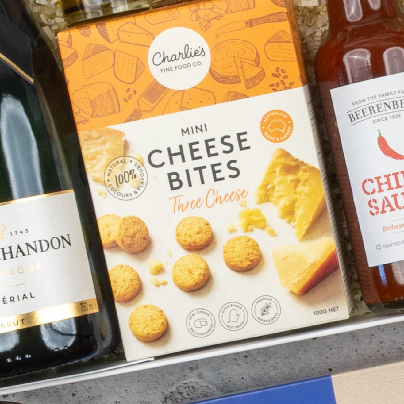 A close-up of a Moët & Chandon Deluxe Celebration Hamper by The Hamper Boutique Co, featuring a box of Charlies Mini Cheese Bites (Three Cheese flavor) between champagne and Berenberg Chilli Sauce.