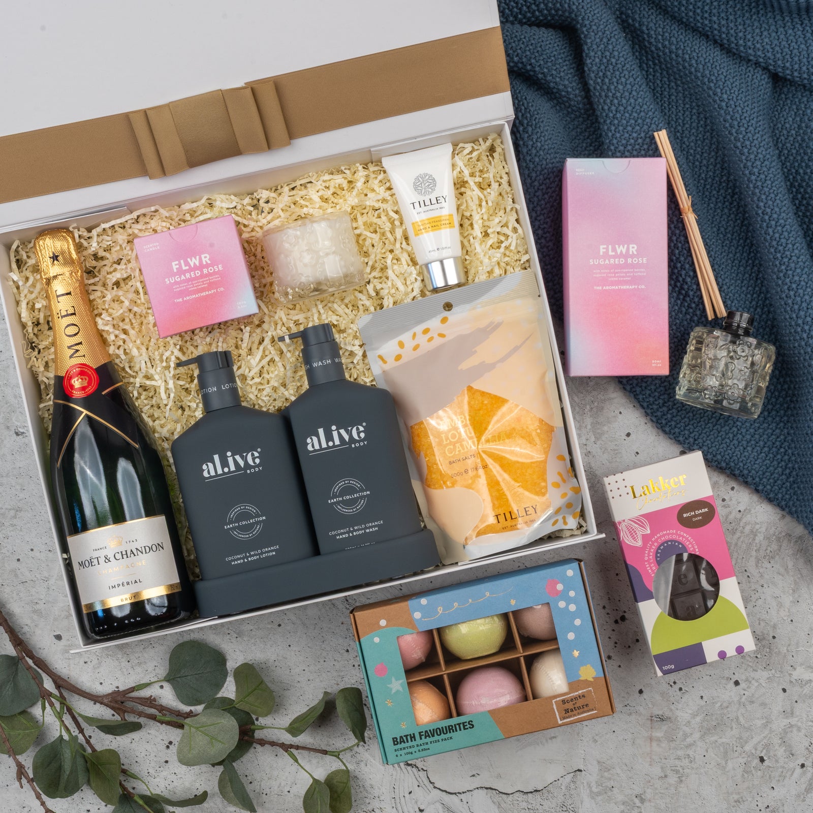 The Hamper Boutique Co’s Champagne Wellness Escape features Moët & Chandon, body wash, lotion, hand cream, bath salt, bath bombs, reed diffuser, candle & chocolate egg—beautifully arranged with green leaves on a textured surface.