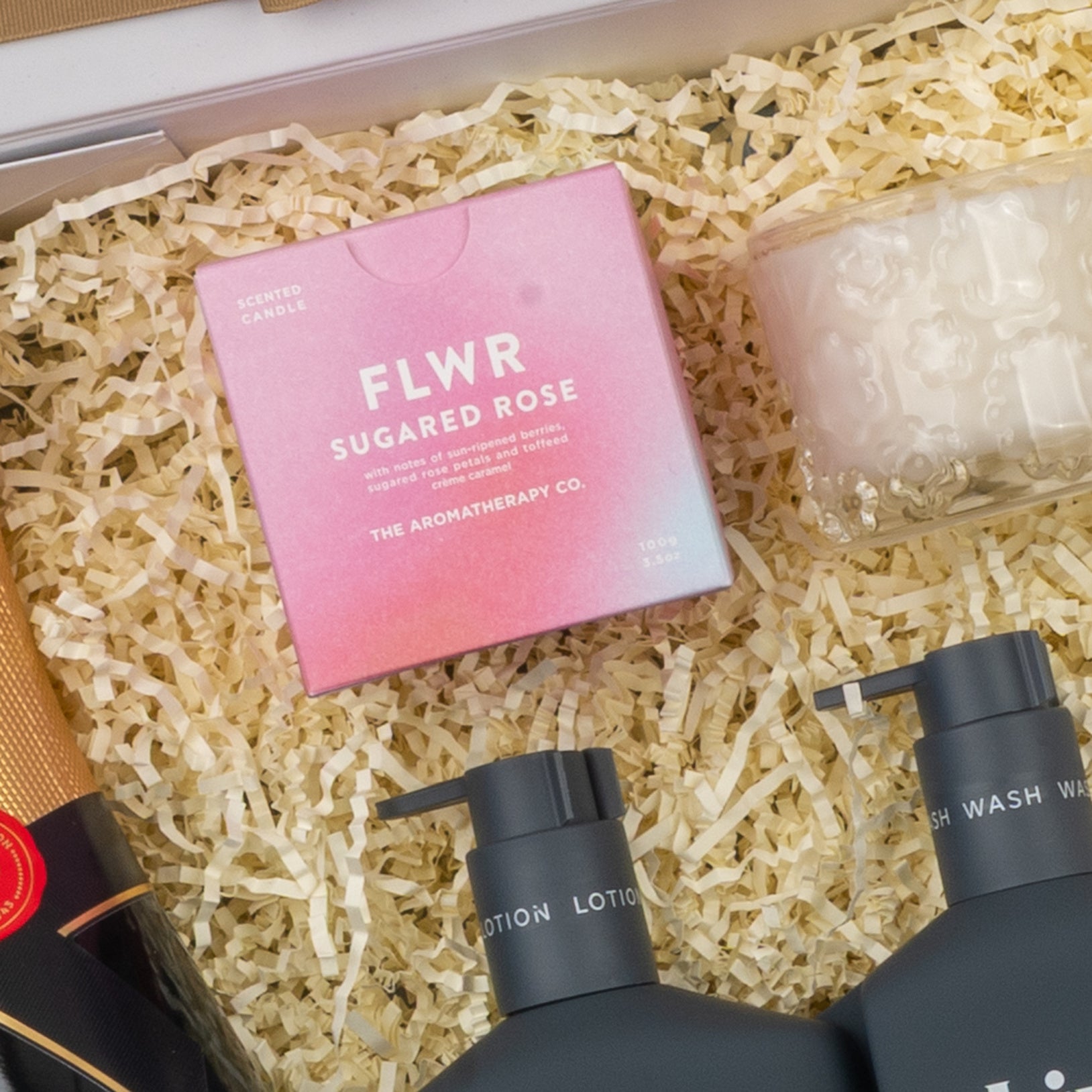 The Hamper Boutique Co's Champagne Wellness Escape gift box includes a pink FLWR Sugared Rose candle, a textured white candle, black bottles of lotion and wash, plus a gold-foil wrapped bottle nestled in shredded paper.