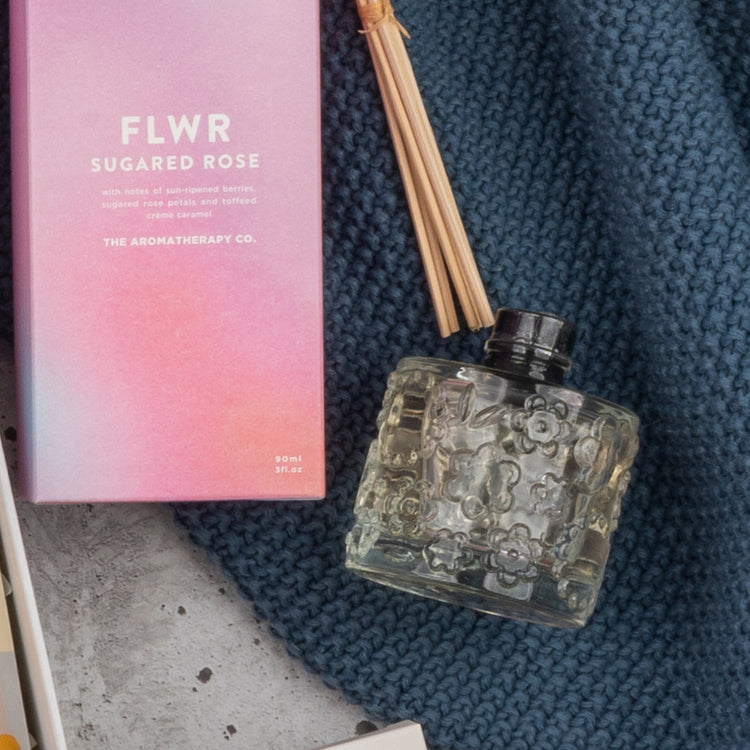 A glass reed diffuser bottle with floral patterns and wooden sticks sits beside a Champagne Wellness Escape box by The Hamper Boutique Co, displayed on blue knitted fabric.