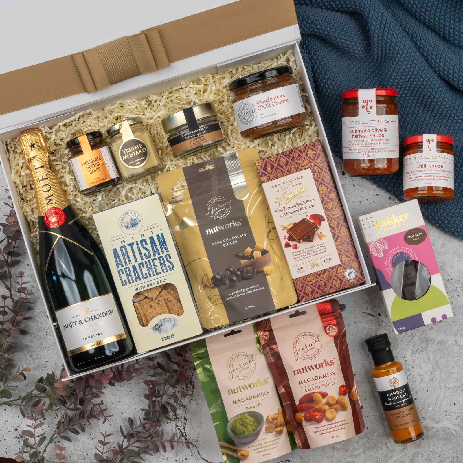 The Hamper Boutique Co’s Moët & Chandon Premium Celebration Hamper features champagne, crackers, chocolates, assorted nuts, cookies, condiments and sauces, beautifully arranged with a blue cloth and decorative leaves.