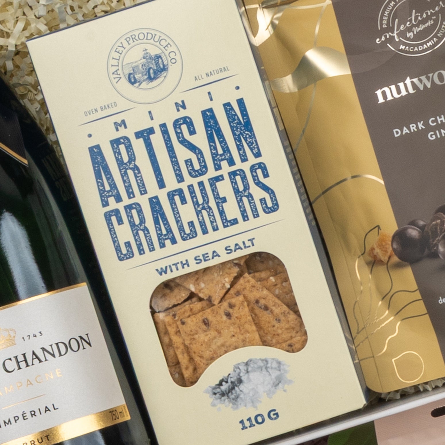 A close-up of The Hamper Boutique Co’s Moët & Chandon Premium Celebration Hamper, featuring mini artisan sea salt crackers, a bottle of Moët & Chandon champagne, and dark chocolate macadamias.