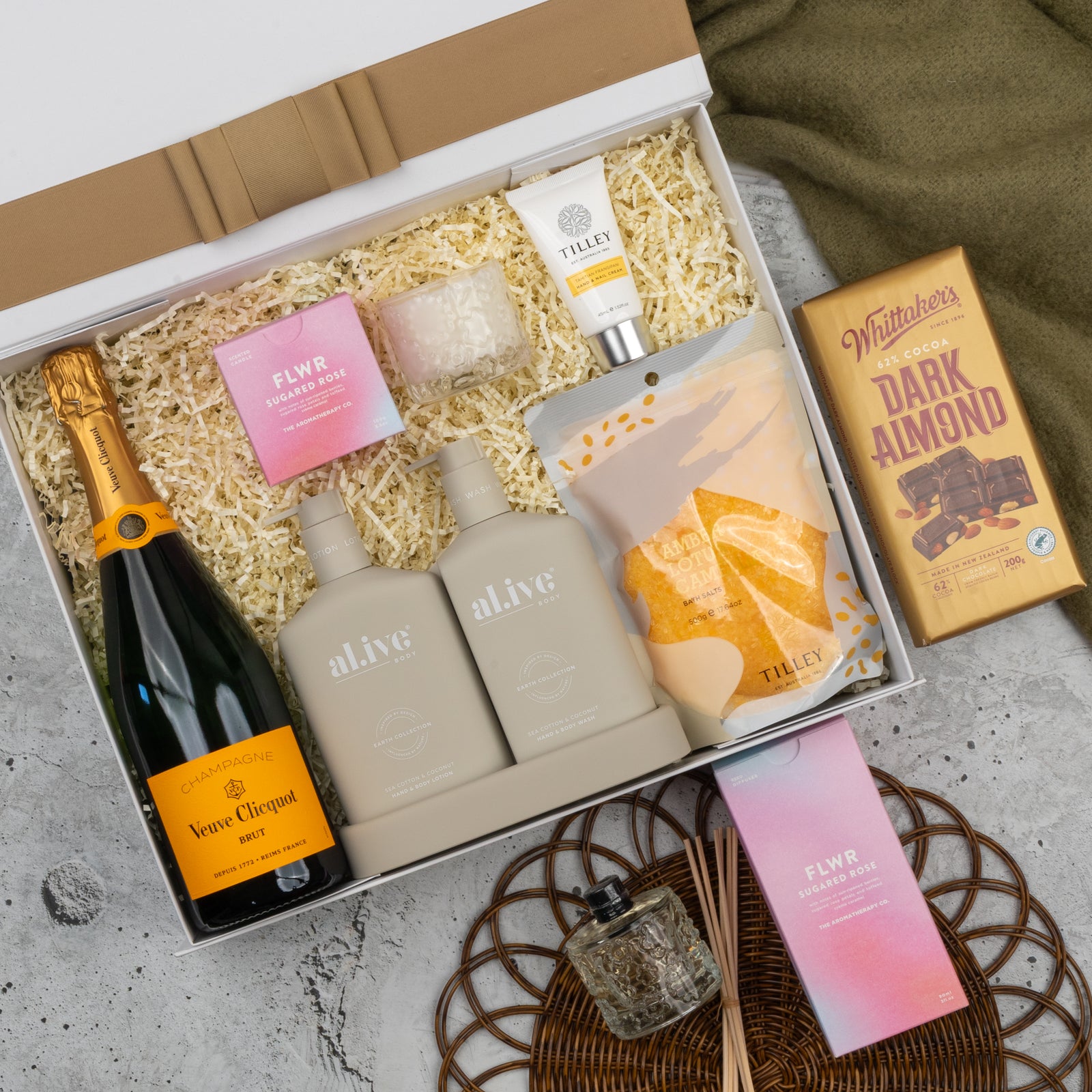 The Hamper Boutique Co’s Veuve Clicquot Premium Celebration Hamper includes champagne, skincare items, two FLWR candles, Tilley lotion, Whittaker’s dark almond chocolate, and a diffuser—beautifully presented with shredded filler.
