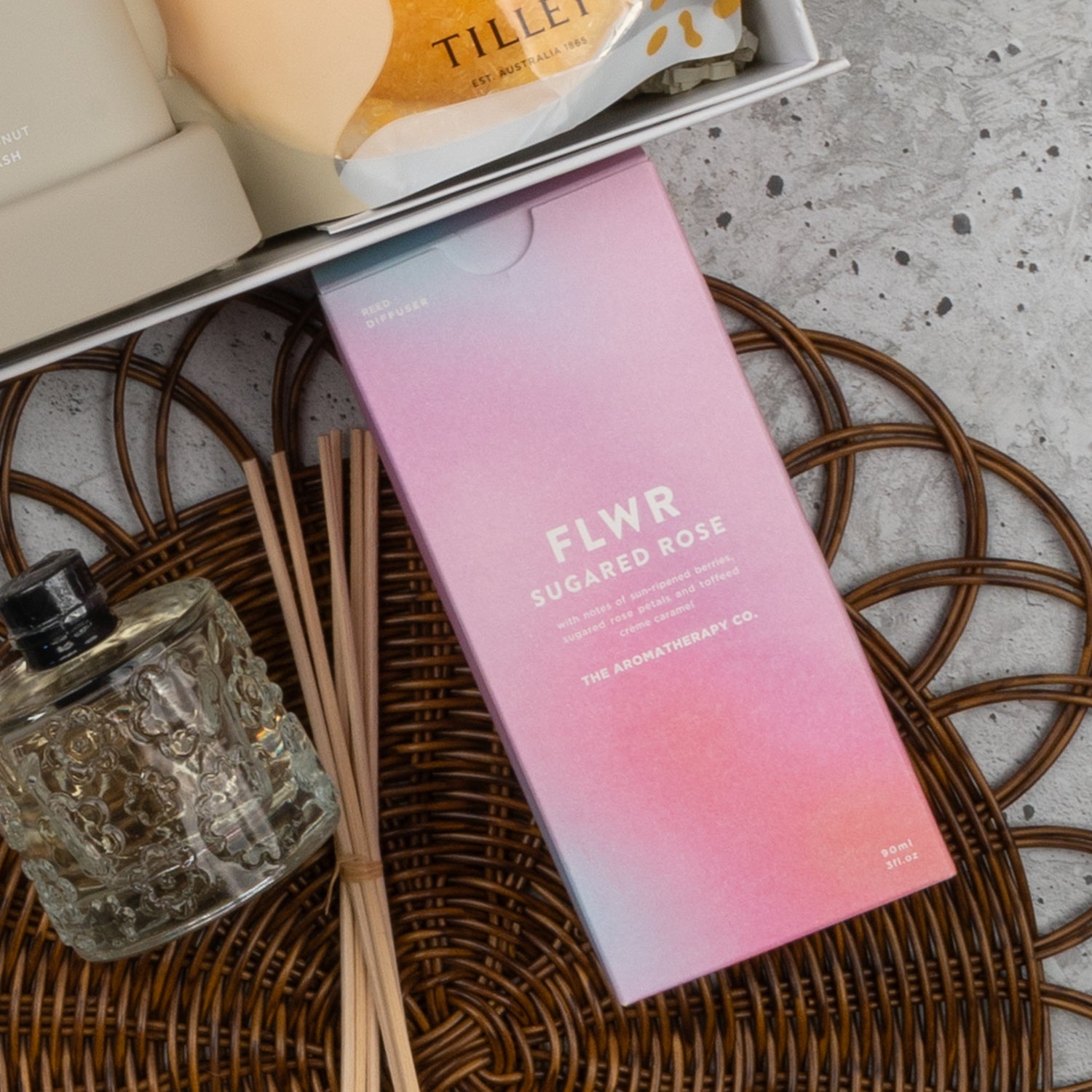 A Veuve Clicquot Premium Celebration Hamper by The Hamper Boutique Co sits on a wicker mat, with its elegant box beside a glass bottle and reed sticks. Other luxury bath items are partially visible in the background.