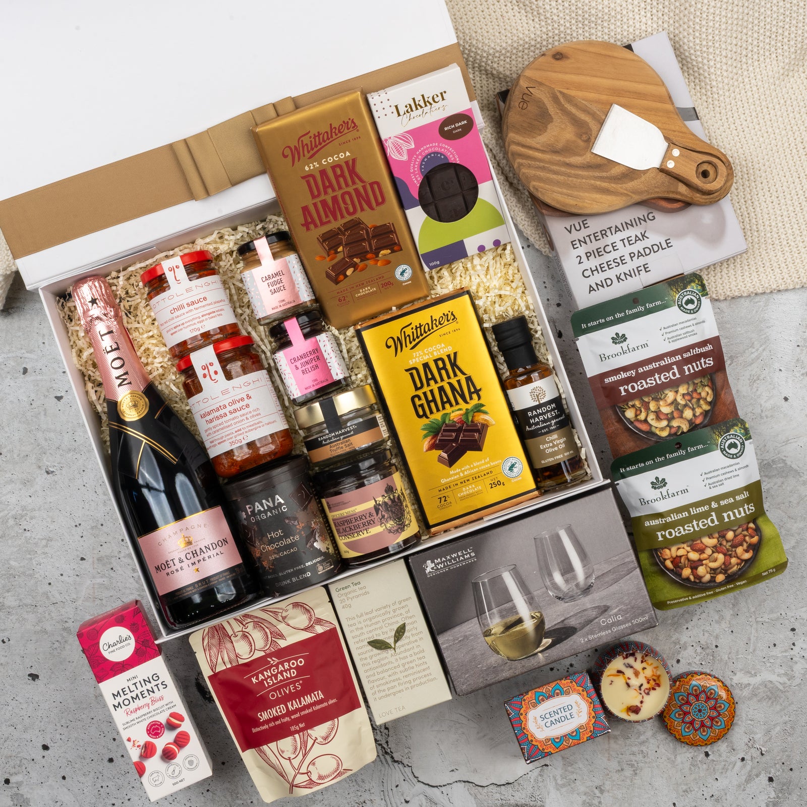 The Hamper Boutique Co's Moët & Chandon Rosé Grand Celebration Hamper features champagne, chocolates, jams, crackers, nuts, a cheese board with knife, wine glasses, and assorted treats, all beautifully arranged on a concrete surface.