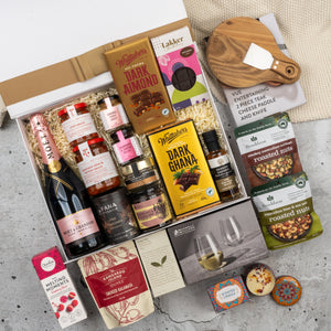 The Hamper Boutique Co's Moët & Chandon Rosé Grand Celebration Hamper features champagne, chocolates, jams, crackers, nuts, a cheese board with knife, wine glasses, and assorted treats, all beautifully arranged on a concrete surface.