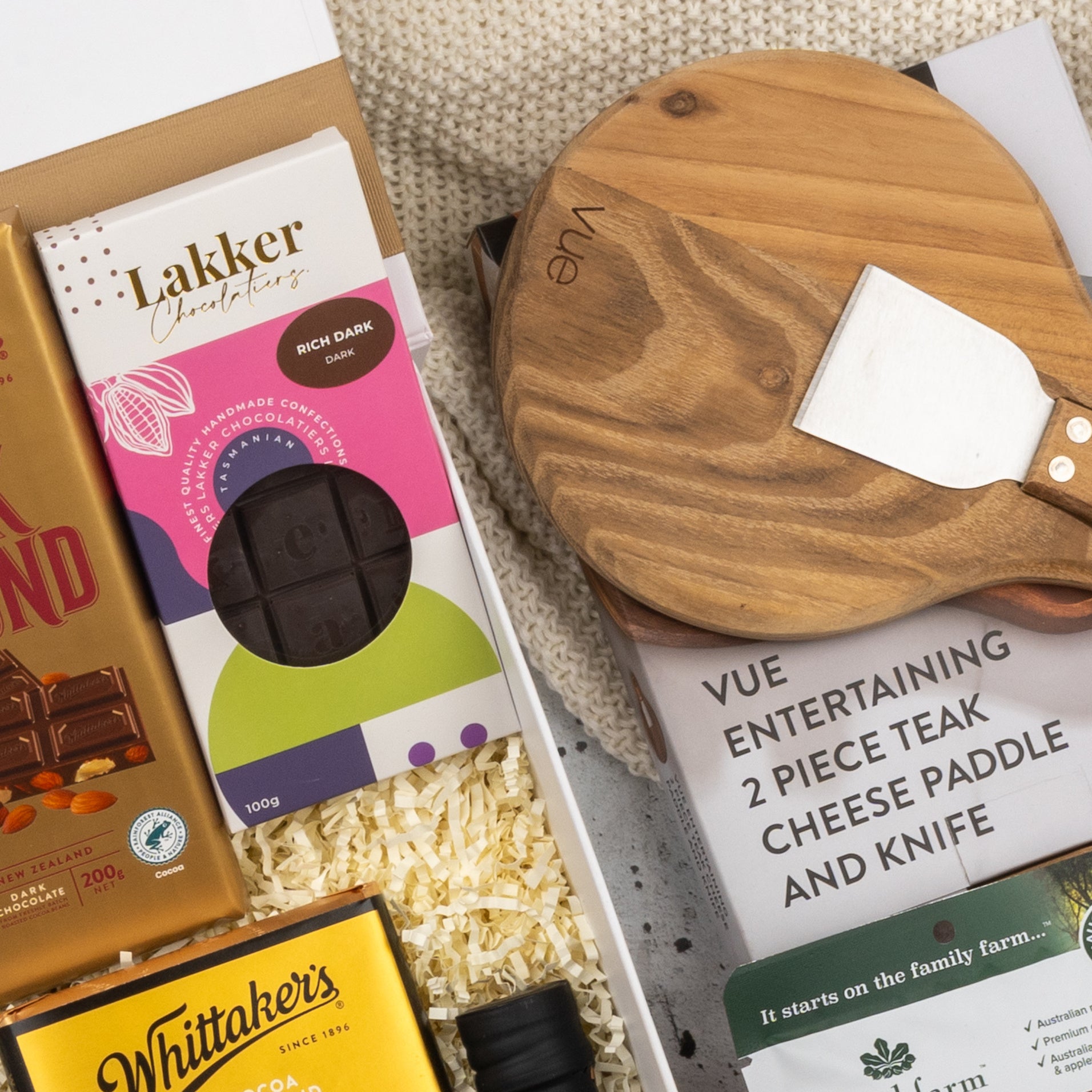 The Hamper Boutique Co’s Moët & Chandon Rosé Grand Celebration Hamper features chocolate bars, a dark chocolate block, a small jar, and a wooden cheese paddle with matching knife, all presented on a knit fabric background.