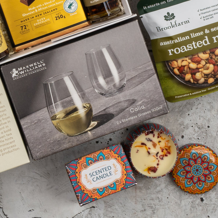 The Moët & Chandon Rosé Grand Celebration Hamper by The Hamper Boutique Co features stemless wine glasses, a scented candle, roasted mixed nuts, artisan chocolate, and a decorative candle, all elegantly arranged on a gray surface.