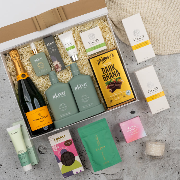 The Hamper Boutique Co's Veuve Clicquot Grand Celebration Hamper includes champagne, two green bottles, chocolate bars, beauty products, candles, and skincare items arranged on crinkle paper atop a textured gray surface.