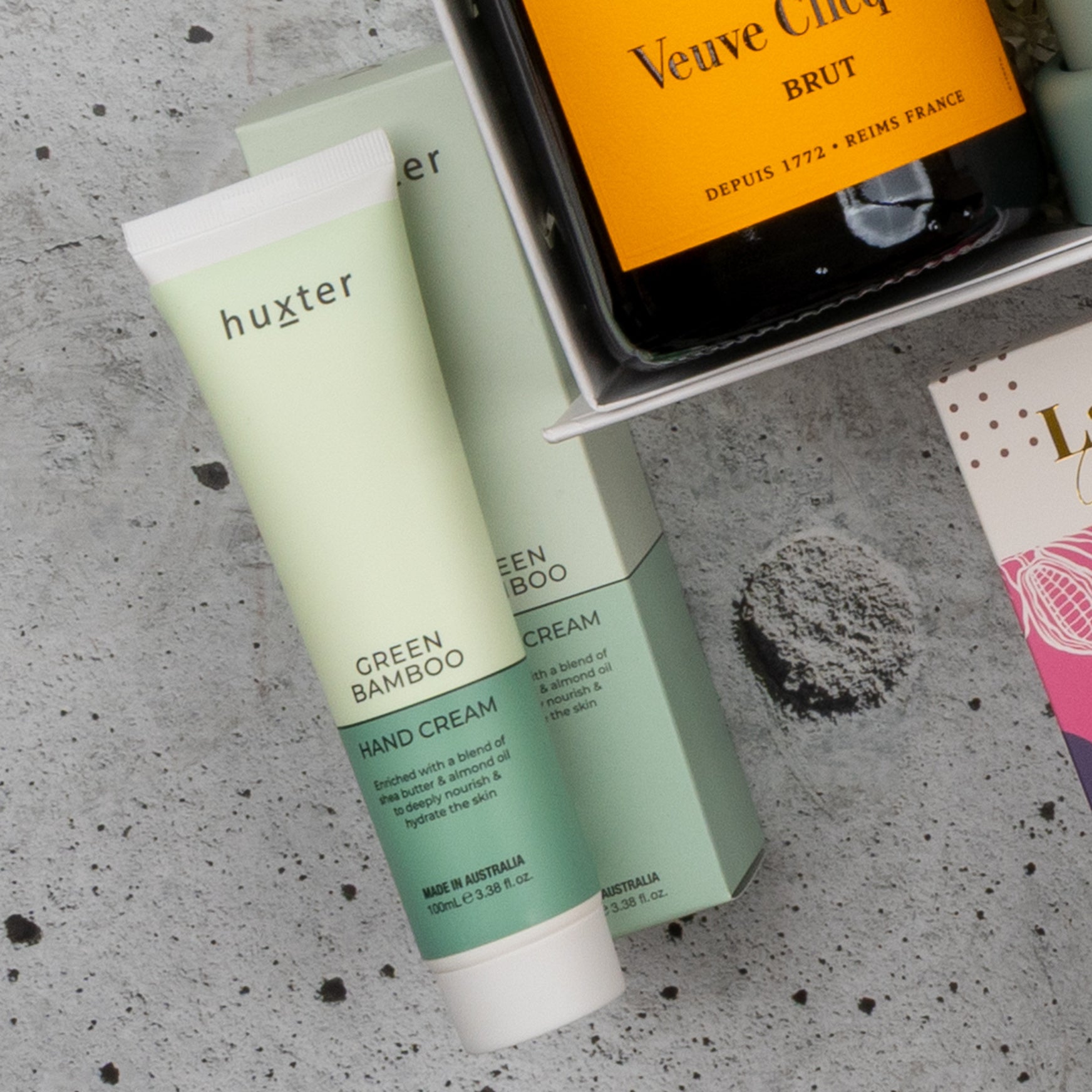 A tube of Huxter Green Bamboo hand cream sits beside its box, with part of a Veuve Clicquot Grand Celebration Hamper by The Hamper Boutique Co and a gift box displayed on a speckled gray surface.