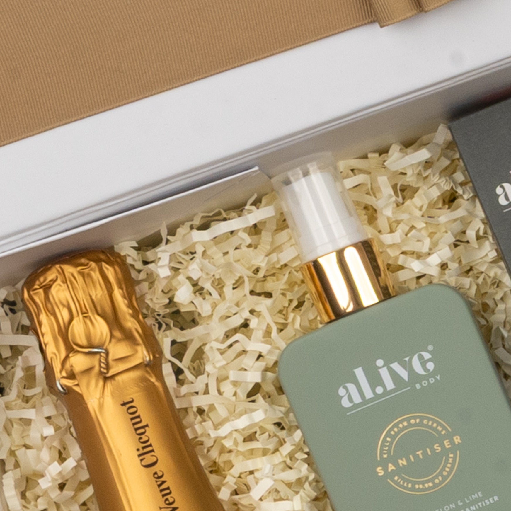 A close-up of The Hamper Boutique Co Veuve Clicquot Grand Celebration Hamper, featuring a gold-wrapped champagne bottle and green al.ive body sanitiser, nestled in shredded paper.