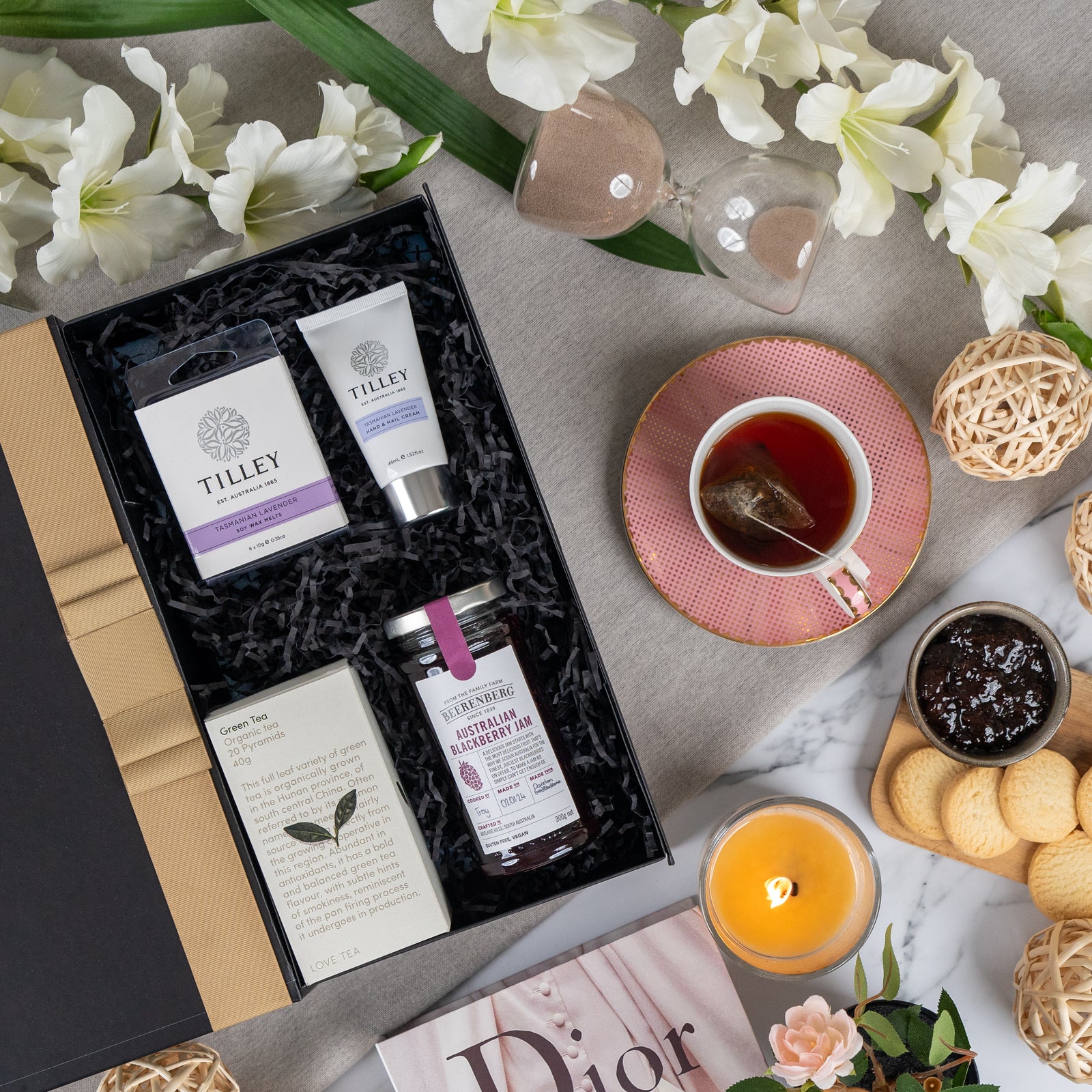 The Hamper Boutique Co’s Self-Care and Relaxation Hamper, featuring hand cream, lotion, tea, and berry conserve, is styled with white flowers, a pink teacup, cookies, jam, a yellow candle, hourglass, and Dior magazine on a gray surface.