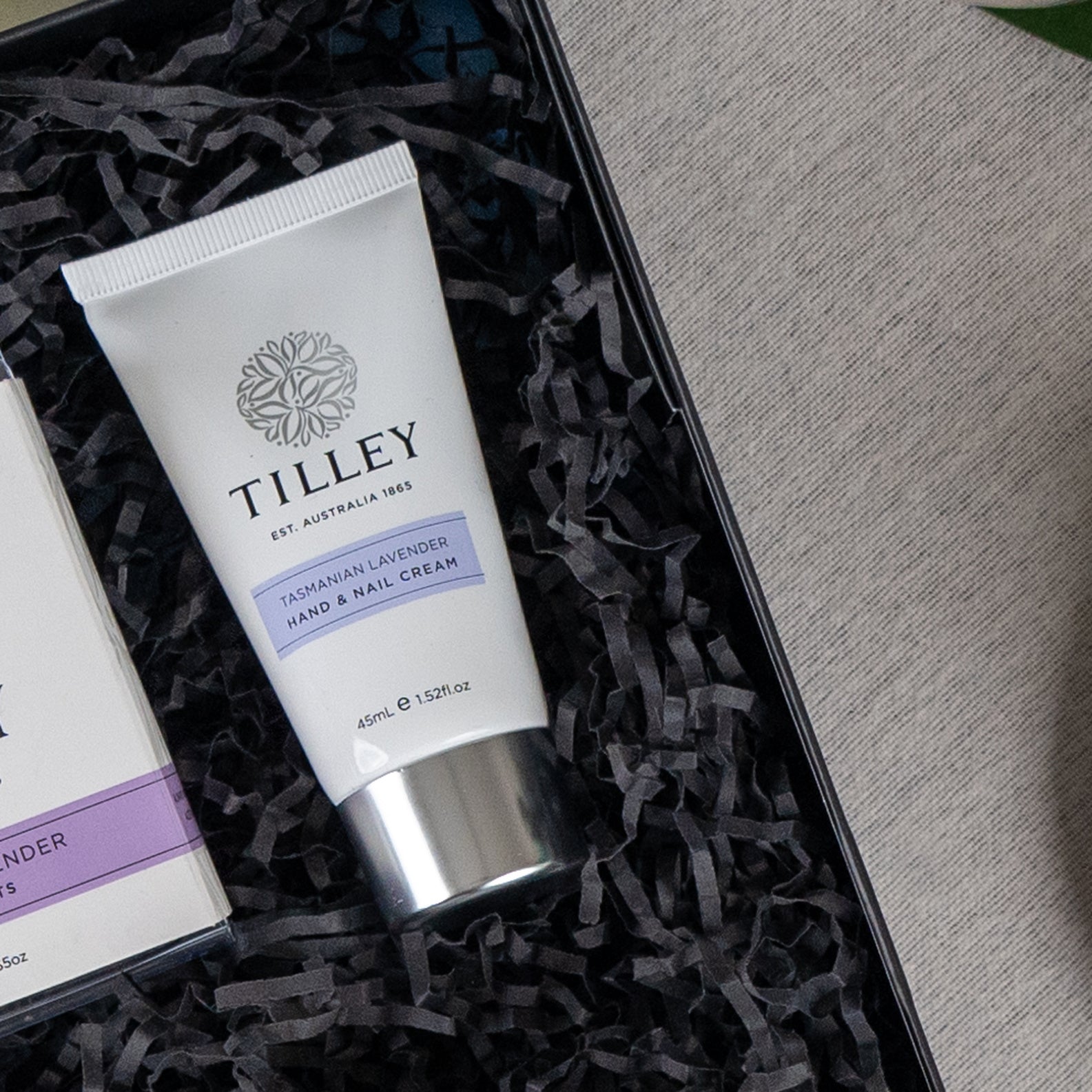 A Self-Care and Relaxation Hamper by The Hamper Boutique Co sits on a grey fabric surface, featuring a gift box filled with black shredded paper and a matching box.