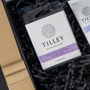 A close-up of The Hamper Boutique Co Self-Care and Relaxation Hamper shows Tilley Tasmanian Lavender soy wax melts with a white and purple label, nestled in black shredded paper beside another Tilley item in a tube.