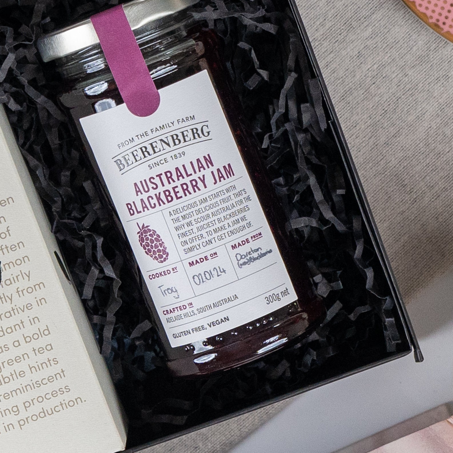 A Self-Care and Relaxation Hamper from The Hamper Boutique Co features a jar of Australian Blackberry Jam nestled in black shredded paper beside an open book, with the jam's label displaying its details and icons.