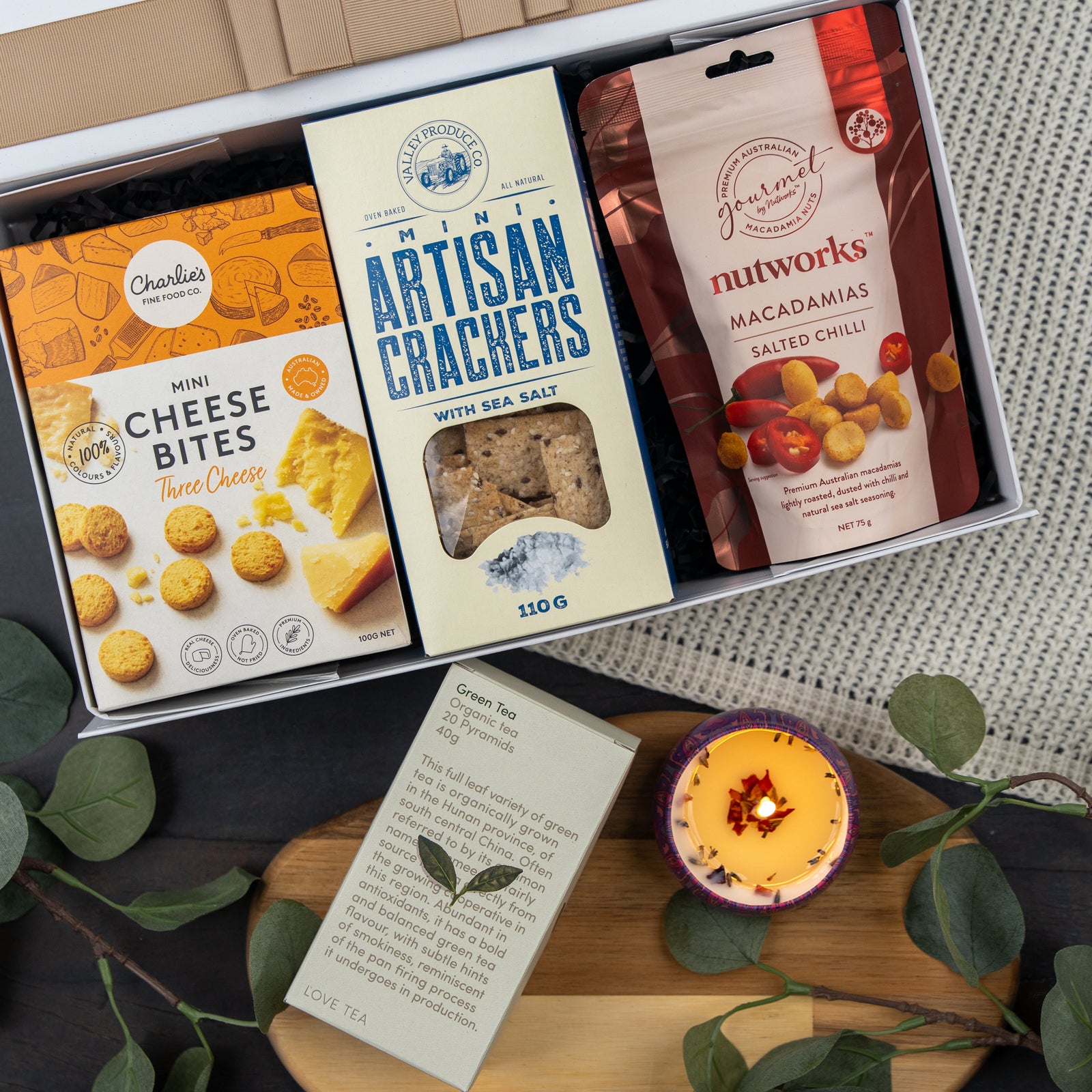The Hamper Boutique Co's Cozy Moments Tea Hamper features mini cheese bites, artisan crackers, salted chili macadamias, a green tea sachet, and a lit candle, beautifully presented with green leaves on a black and gray surface.