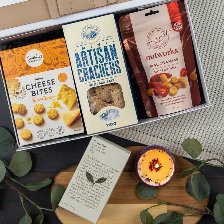 The Hamper Boutique Co's Cozy Moments Tea Hamper features mini cheese bites, artisan crackers, salted chili macadamias, a green tea sachet, and a lit candle, beautifully presented with green leaves on a black and gray surface.
