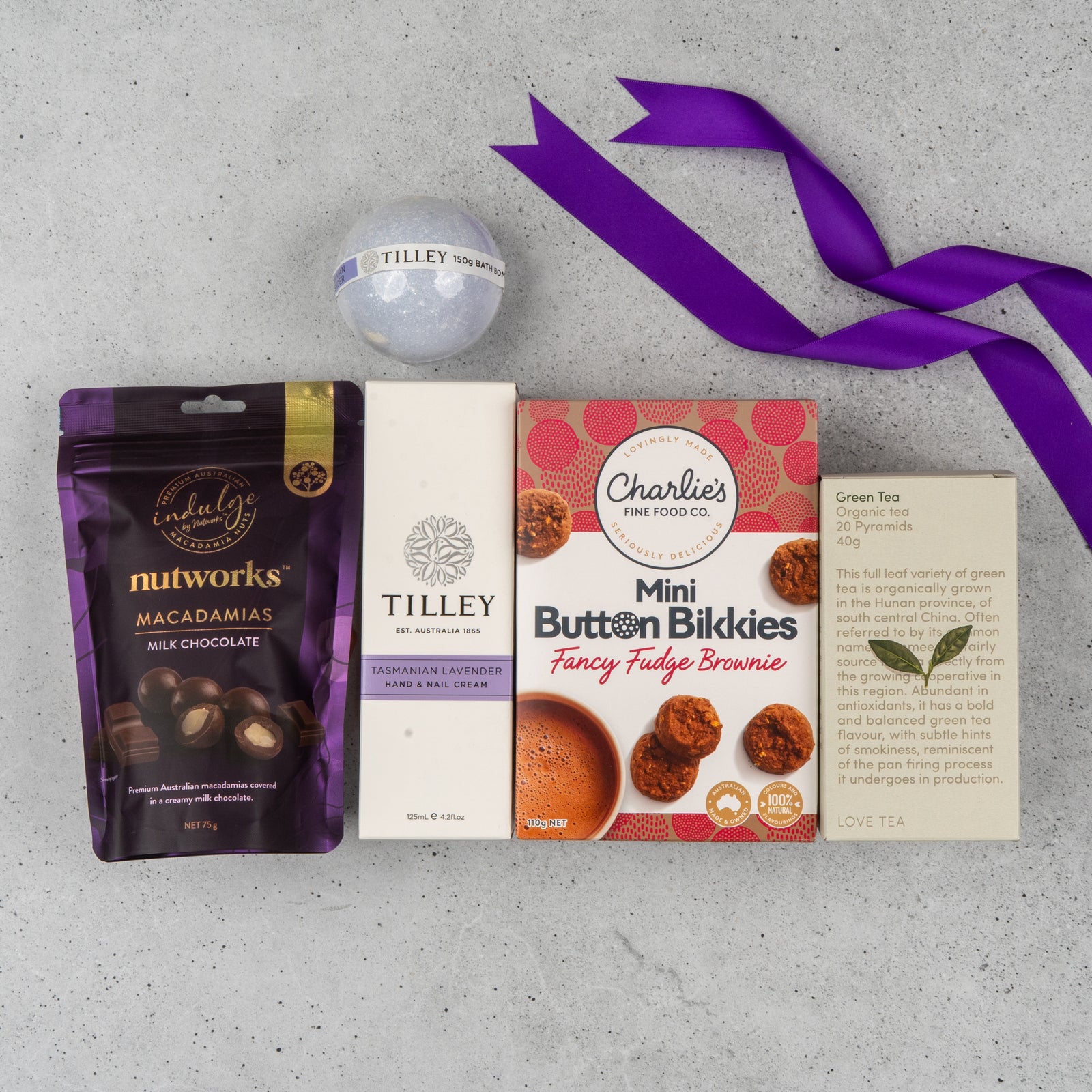 The Hamper Boutique Co's Tea Lover's Essentials Hamper features milk chocolate macadamias, Tilley Tasmanian lavender hand cream, a bath bomb, Charlies Mini Button Bikkies, a green tea card, and is finished with a purple ribbon.