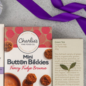 A Tea Lover's Essentials Hamper from The Hamper Boutique Co sits beside a box of Charlies Mini Button Bikkies Fancy Fudge Brownie, both tied with a purple ribbon on a gray surface.