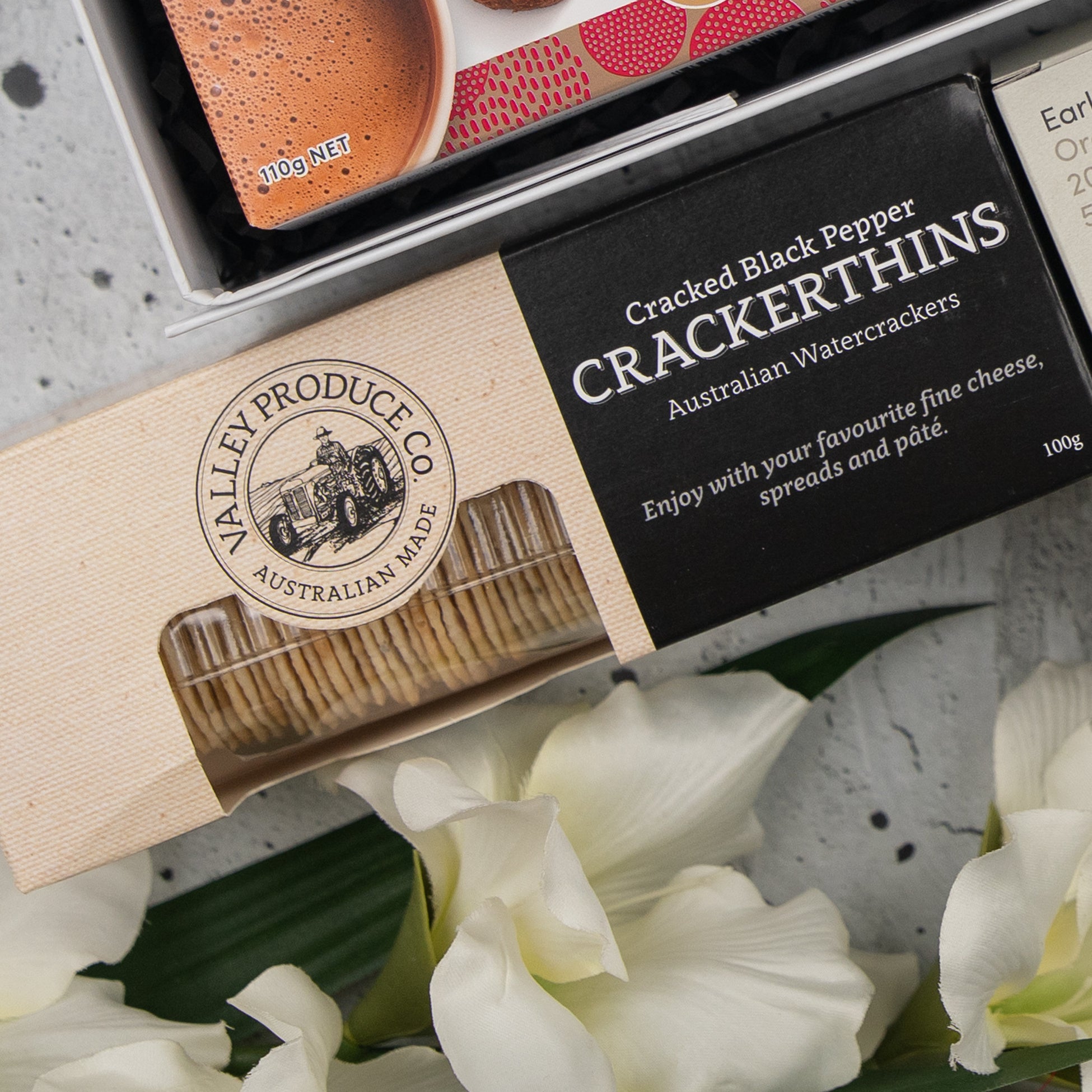 A box of The Hamper Boutique Co Black Tea Haven Hamper sits partially open next to white flowers, with another food box partially visible above.