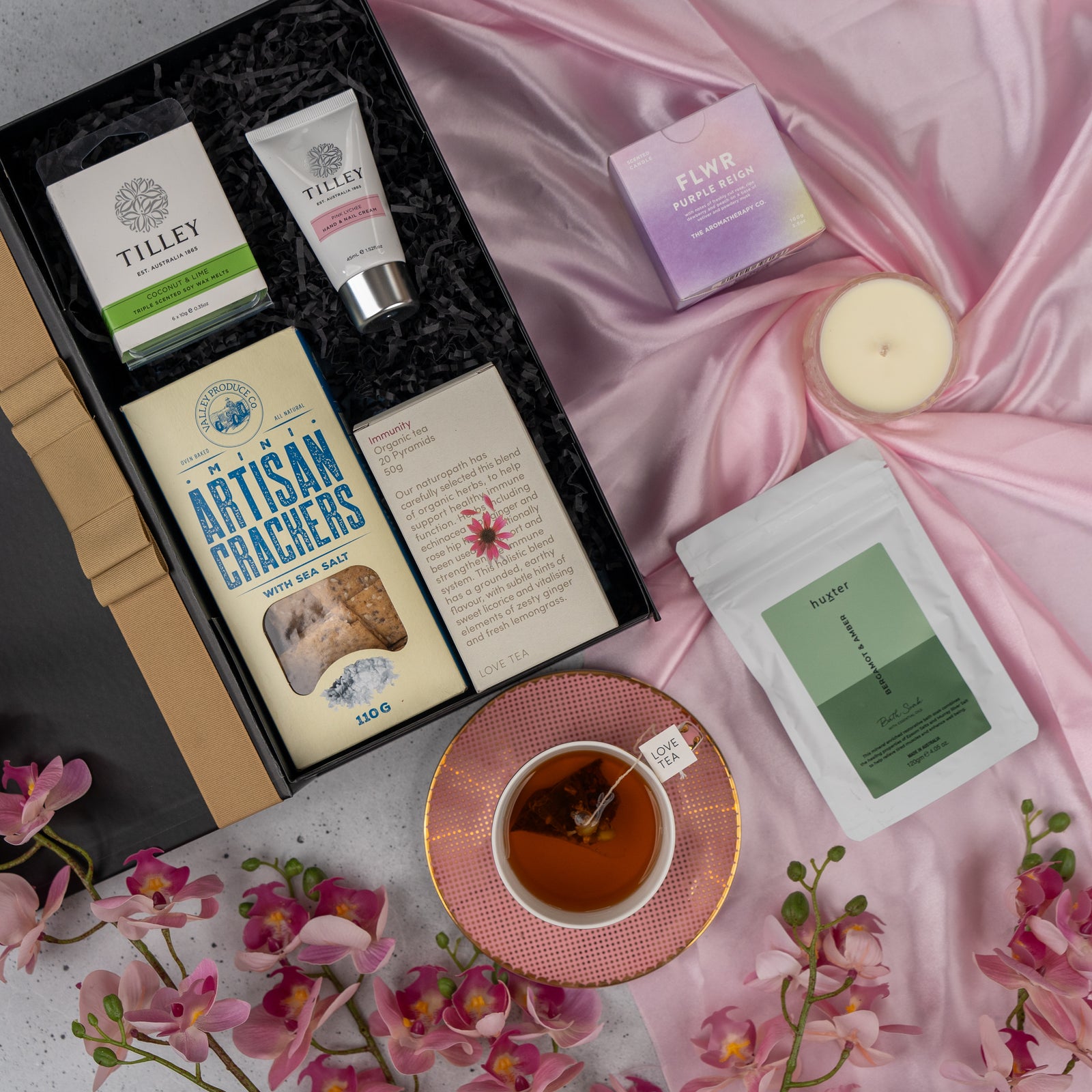 The Hamper Boutique Co’s Home and Wellness Edition Hamper features soap, hand cream, crackers, and tea on pink satin, paired with a candle, tea cup with For You tag, tea pouch, and pink orchids.