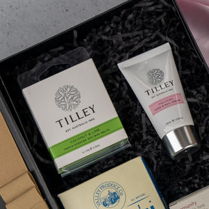 The Hamper Boutique Co Home and Wellness Edition Hamper features Tilley coconut & lime scent melts (white-green box) and Tilley hand & nail cream (white tube, pink label), beautifully arranged on black shredded paper.