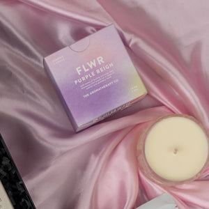 A Home and Wellness Edition Hamper by The Hamper Boutique Co rests on pink satin, featuring a pastel-colored FLWR Purple Reign scented candle beside a beige single-wick candle.
