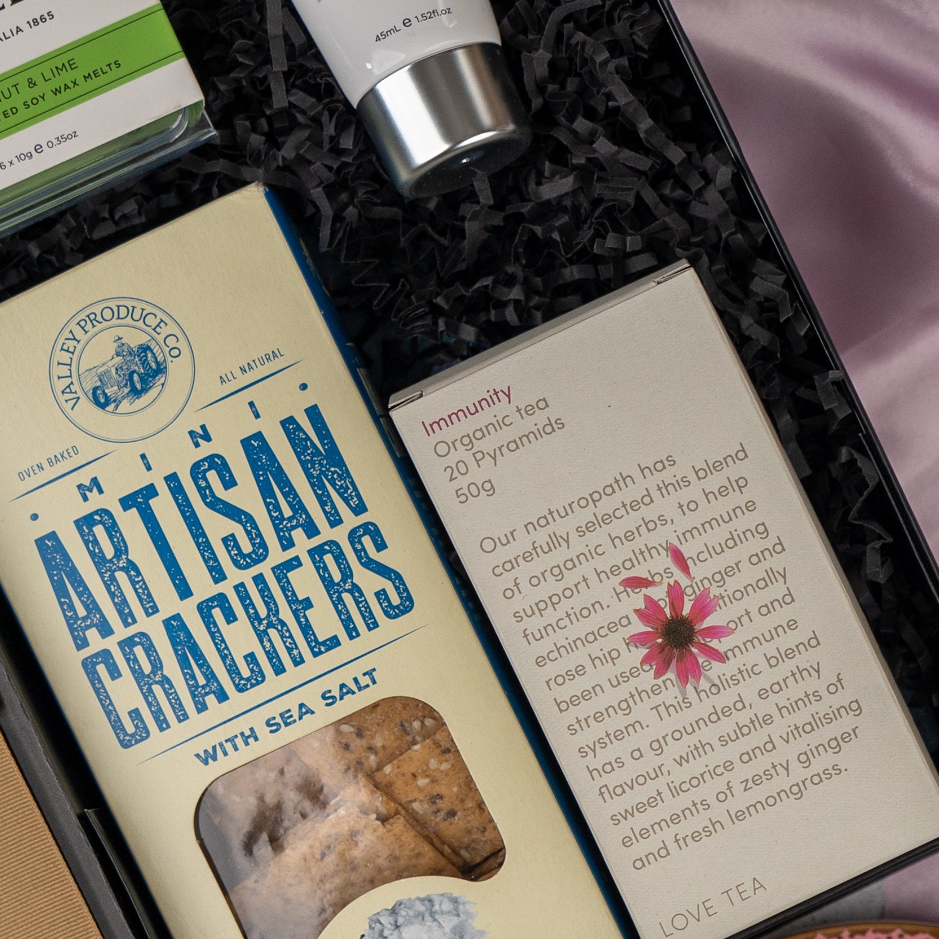A close-up of The Hamper Boutique Co's Home and Wellness Edition Hamper, featuring mini artisan crackers, organic immunity tea, a tube of cream, and a green box, all arranged on black shredded paper.