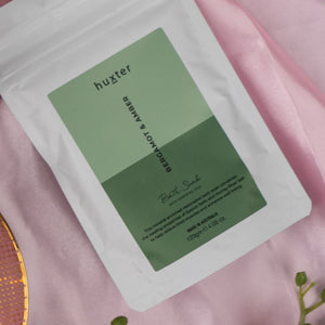 A white resealable pouch from The Hamper Boutique Co’s Home and Wellness Edition Hamper sits on pink fabric, alongside a gold object and green plant. The packaging displays green color blocks and black text.