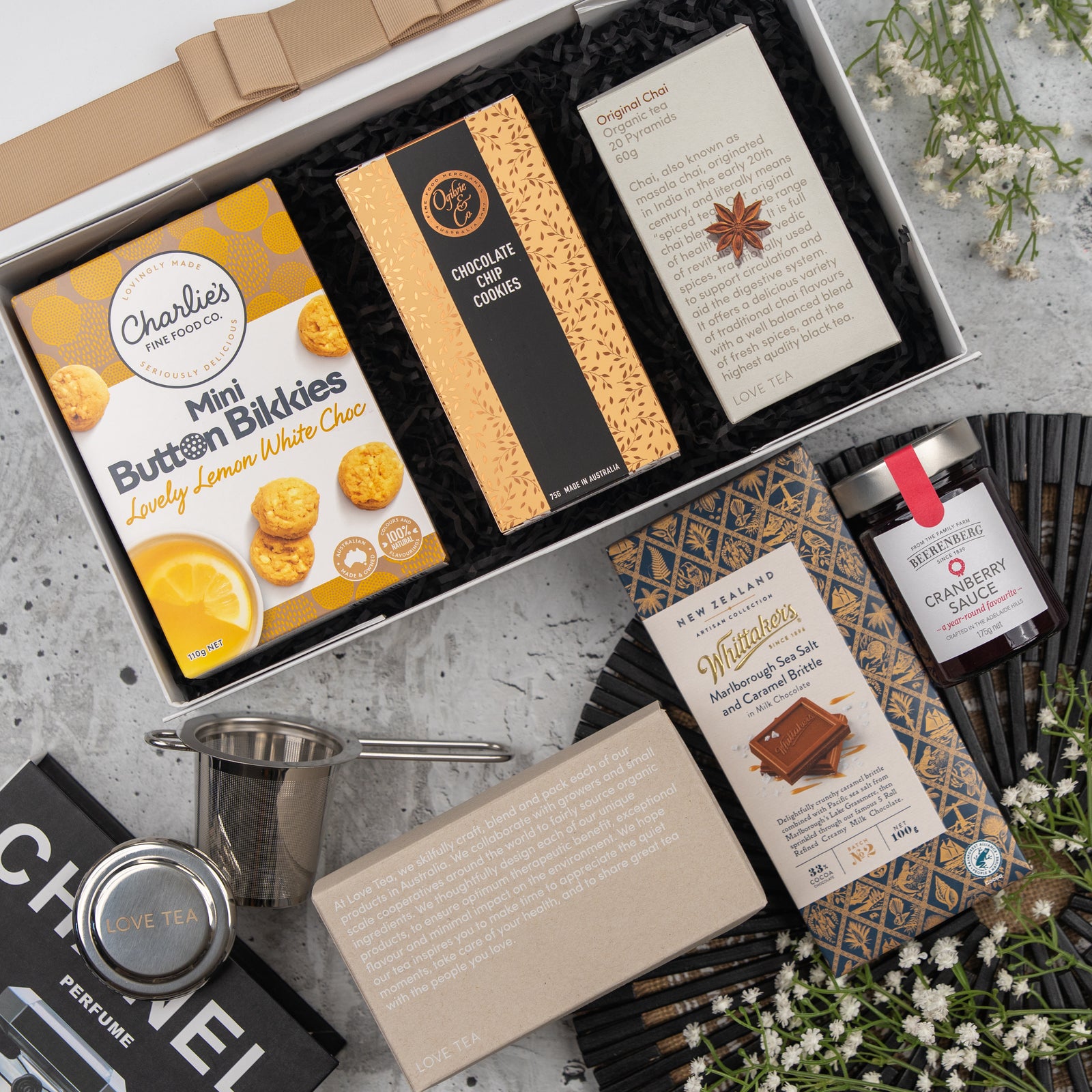 The Exotic Explorations Tea Hamper by The Hamper Boutique Co features mini lemon white chocolate cookies, chocolate chip cookies, chai, cranberry sauce, dark chocolate, a tea infuser, and a tea box arranged on a light floral background.